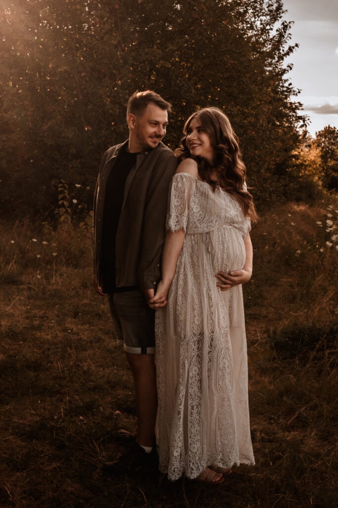 Schwangere Frau in einem fließenden weißen Kleid bei Sonnenuntergang in der Natur nahe Weißwasser – stilvolles Babybauchshooting.