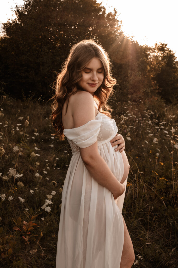 Natürliches Babybauchshooting im Sonnenuntergang – eine schwangere Frau in weißem Kleid auf einer blühenden Wiese in Hoyerswerda, fotografiert von Familienfotografin Elisa Witschas.
